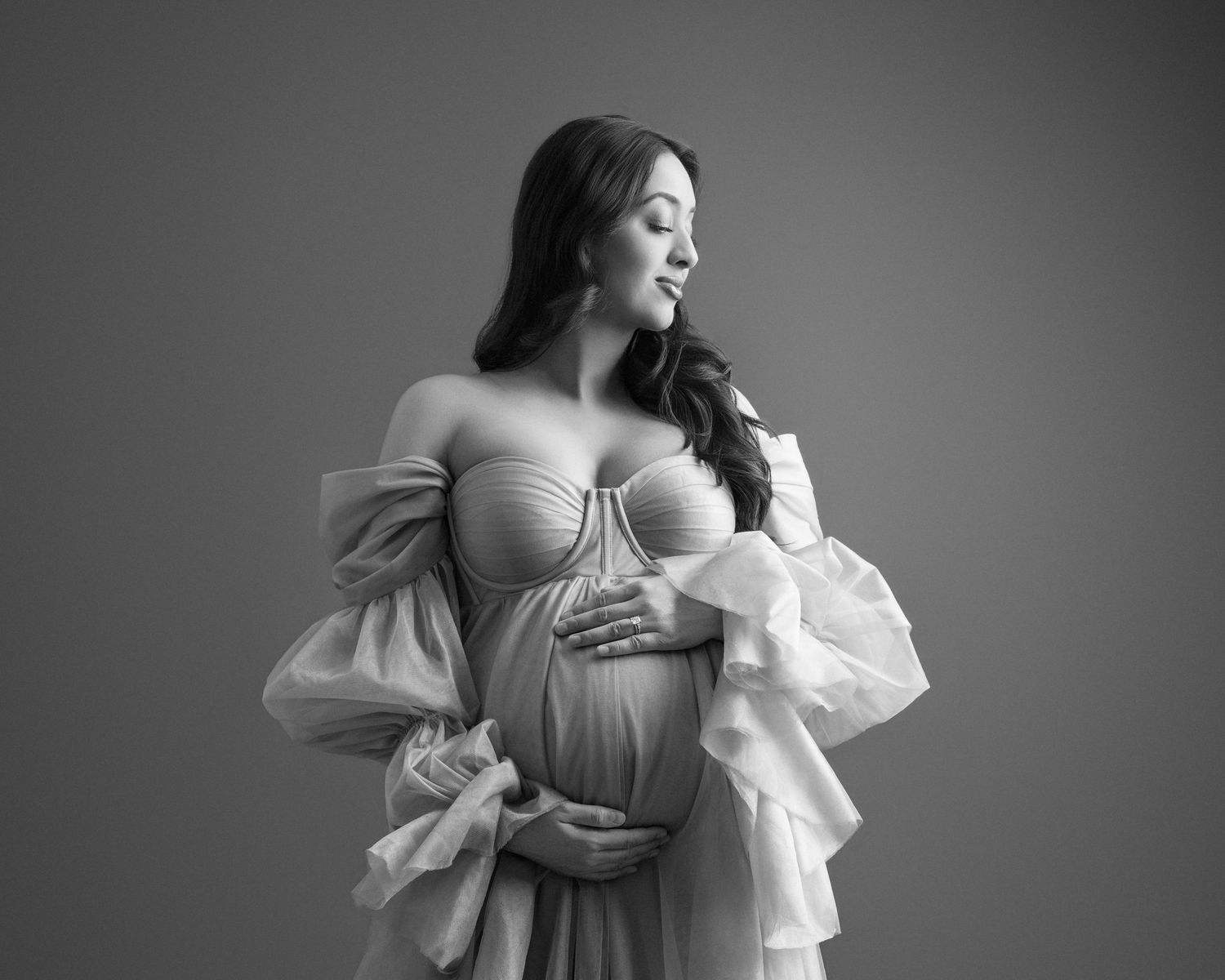 Black and white maternity photo of a pregnant woman in a flowing gown, cradling her belly in a studio setting.