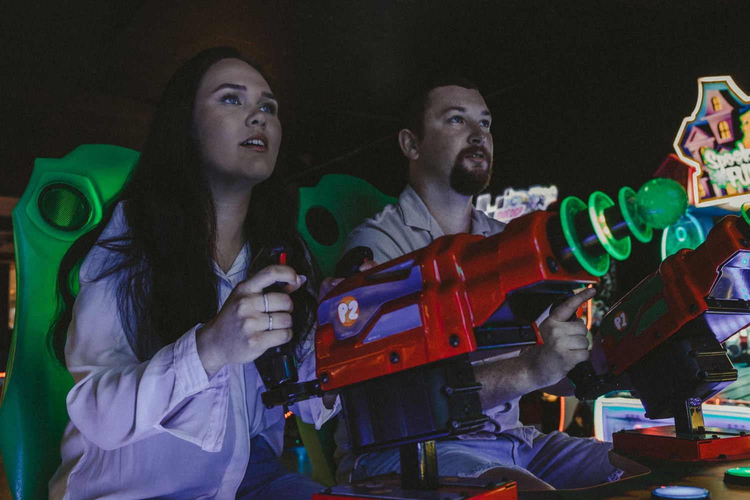 Two people holding colorful toy guns play an arcade shooting game in a dark, vibrant amusement center.