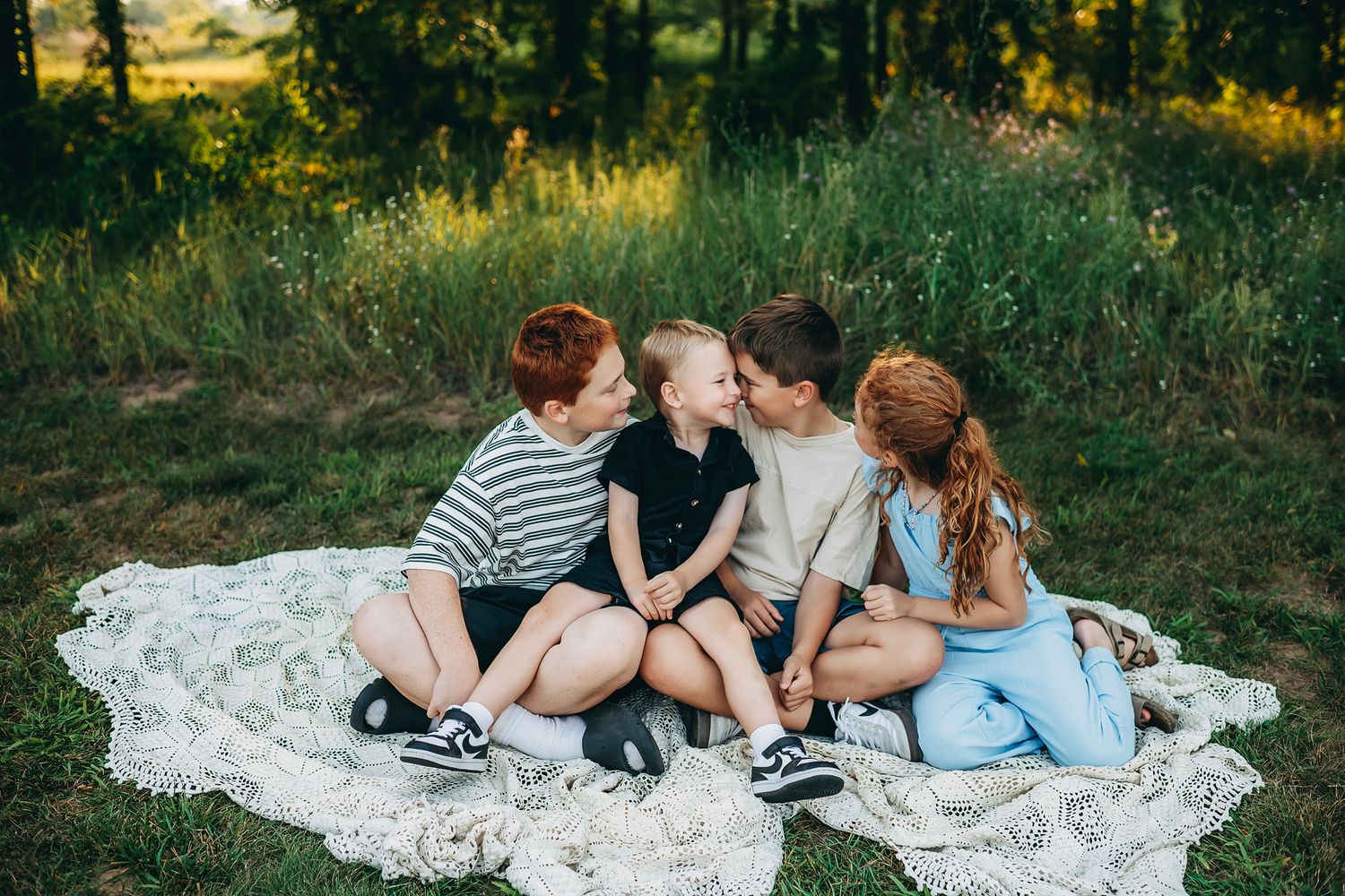 Four children sitting together on a lace blanket outdoors, laughing and hugging in a green meadow.