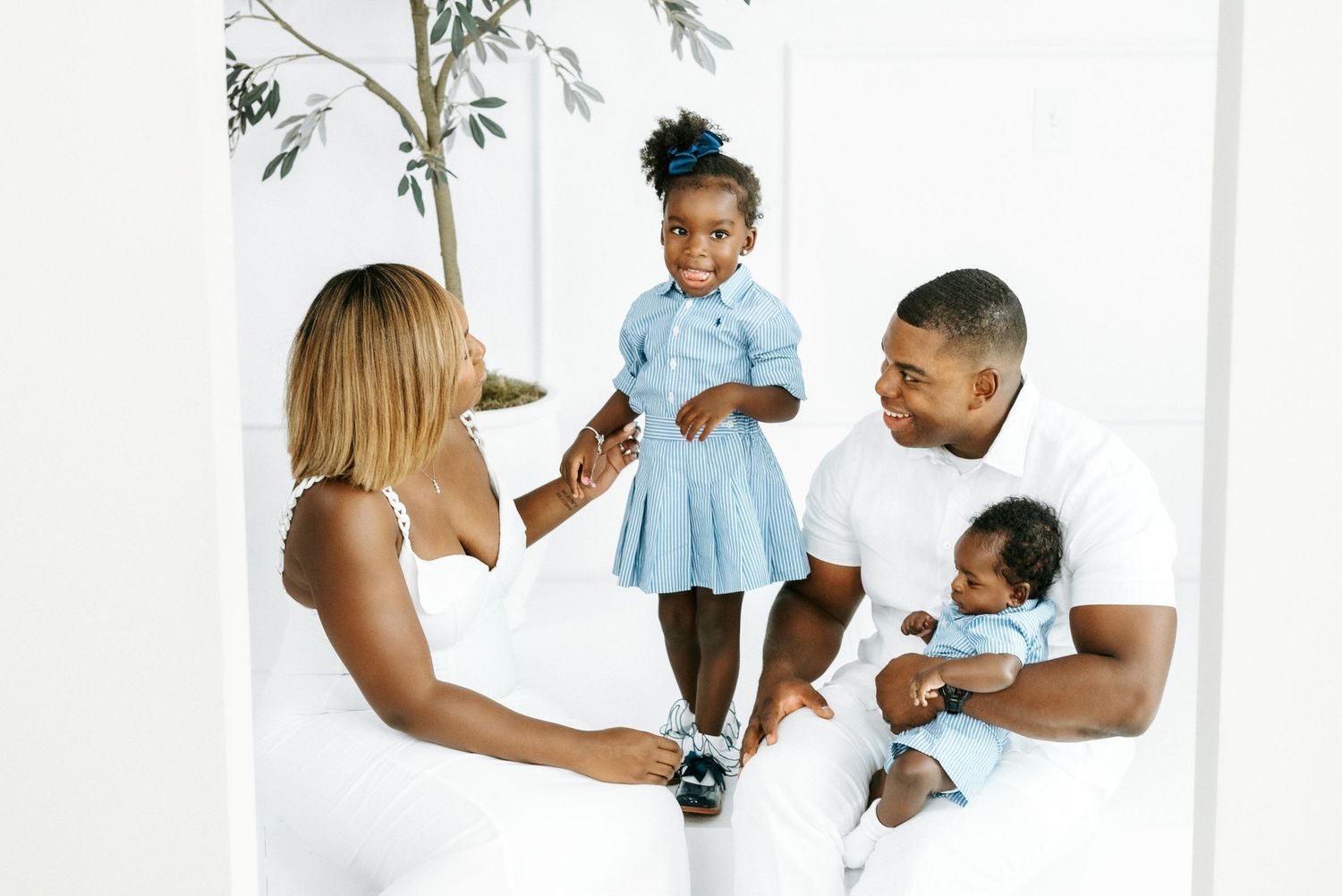 Happy Black family in white outfits with toddler girl in blue dress and newborn baby in bright studio.