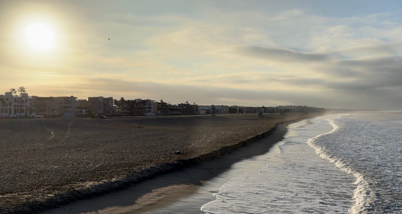 A panoramic view of a sandy beach coastline with rolling waves and a glowing sun in the cloudy sky.