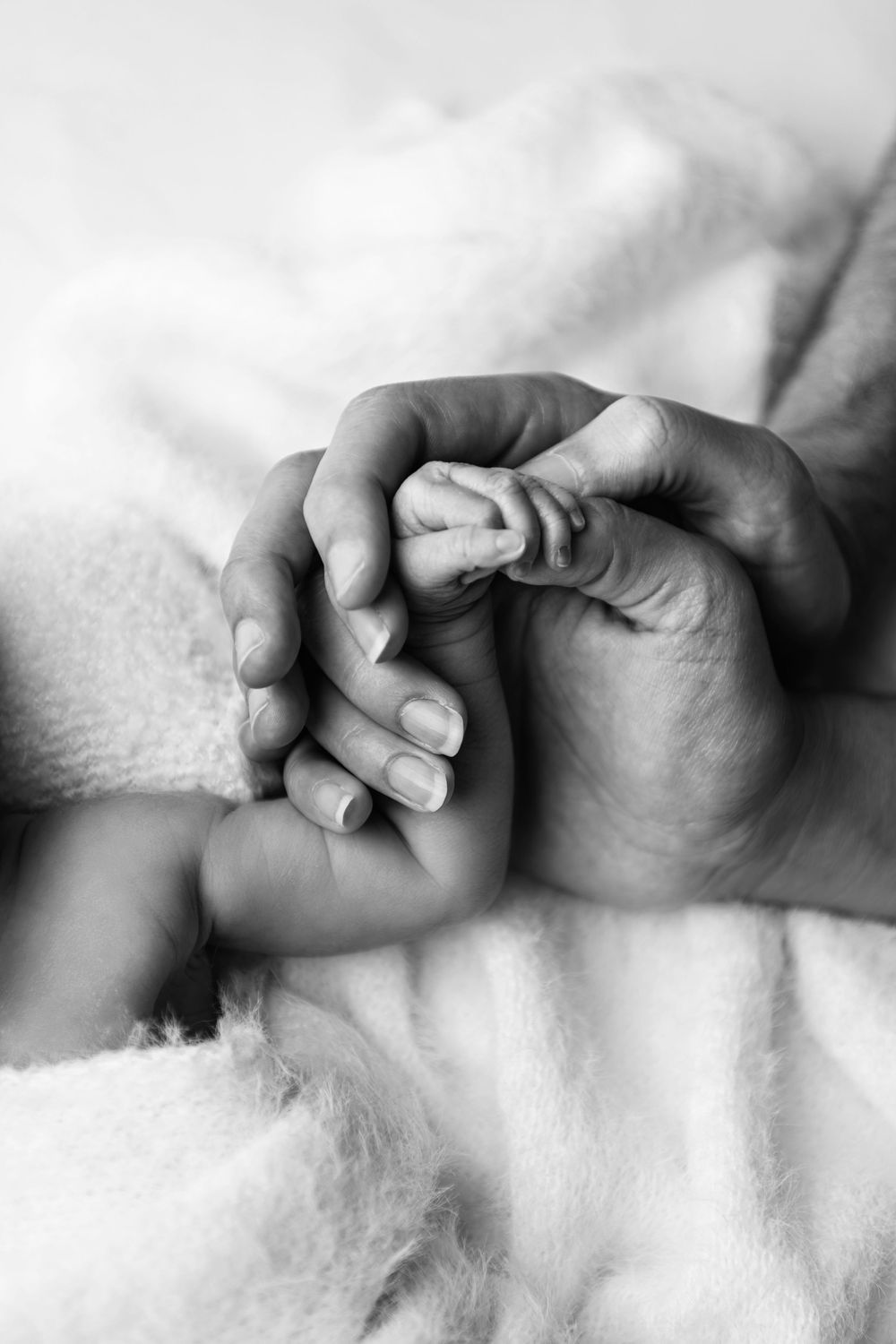 Black and white photo of a newborn baby grasping an adult's finger on a soft white blanket.