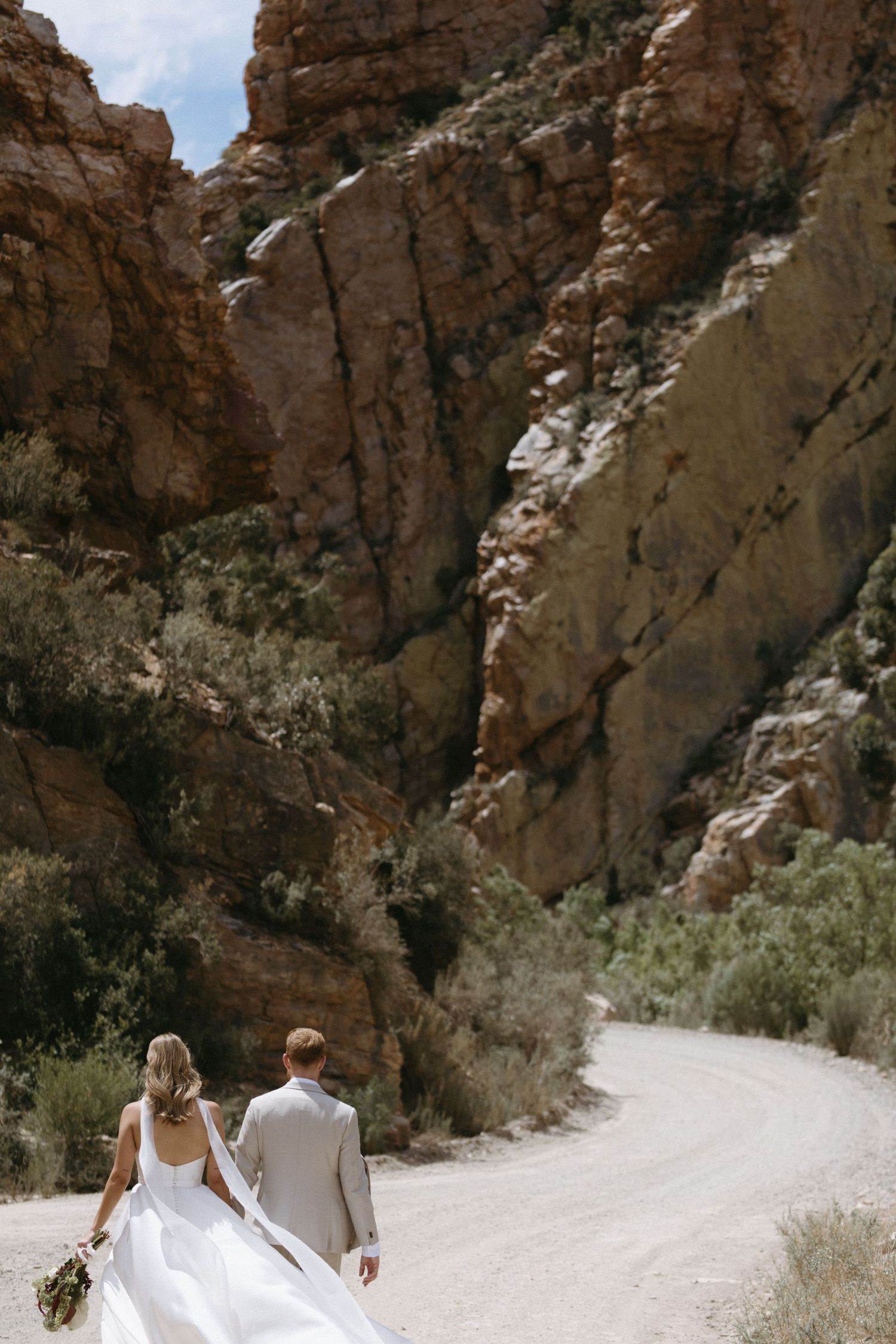 Bride and groom walking hand in hand down a dirt path surrounded by dramatic red rock canyon walls on their wedding day.
