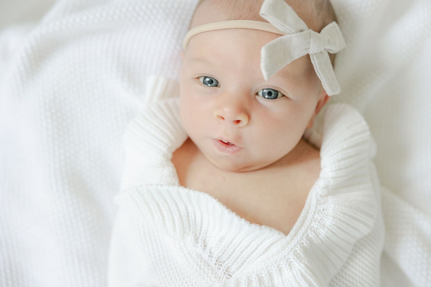 Sweet baby in white knit sweater and bow headband lying on soft white blanket with bright blue eyes. CT Newborn Photography