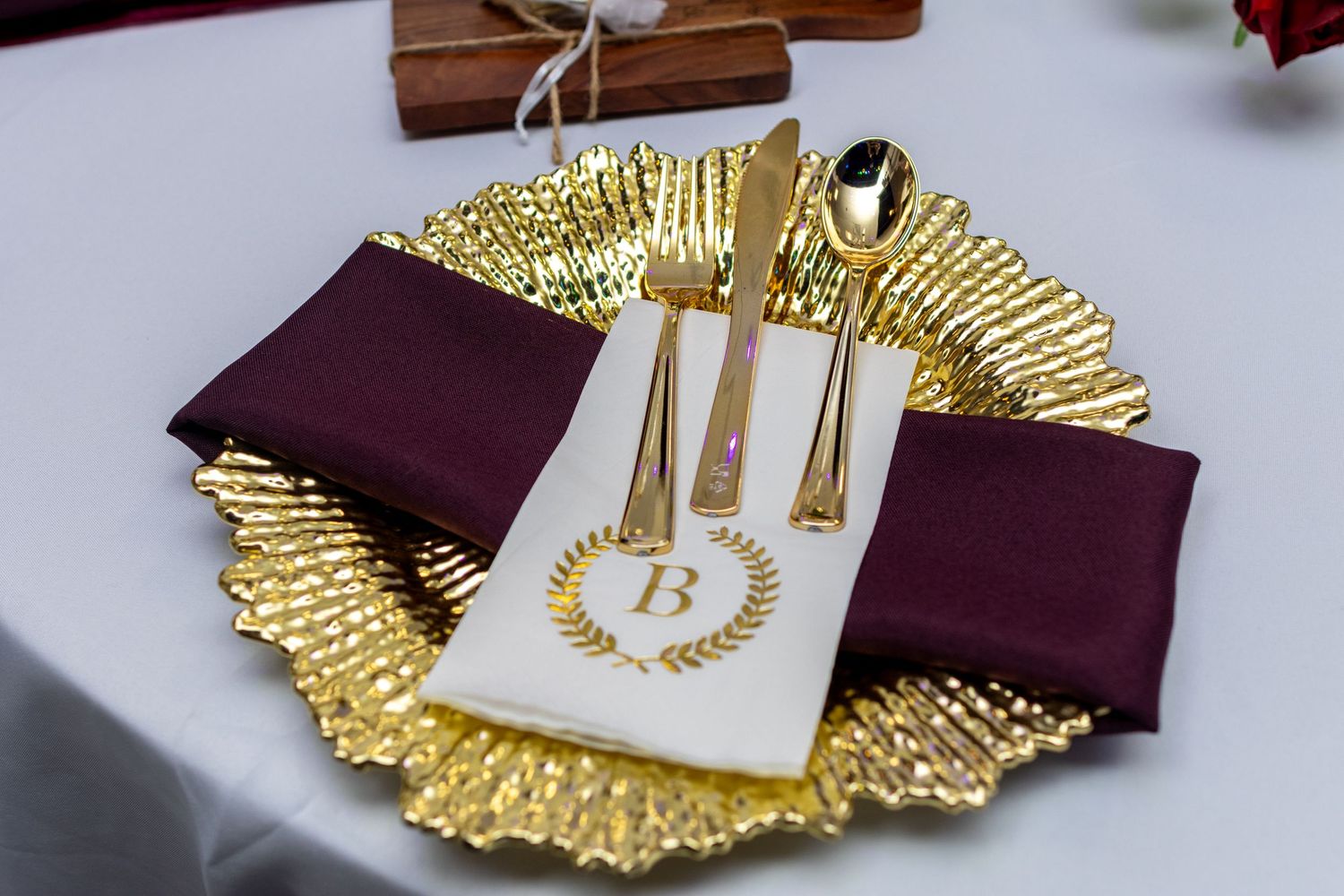 Elegant gold and burgundy place settings with metallic charger plates and utensils arranged on white tablecloth.