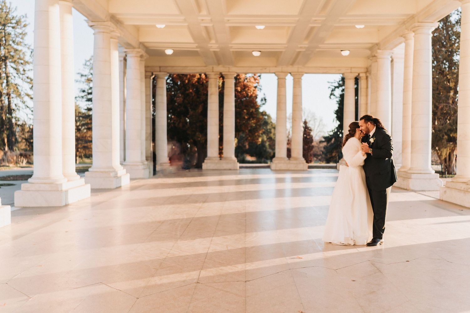 A couple shares a romantic moment in an elegant white columned colonnade with soft lighting and natural surroundings.