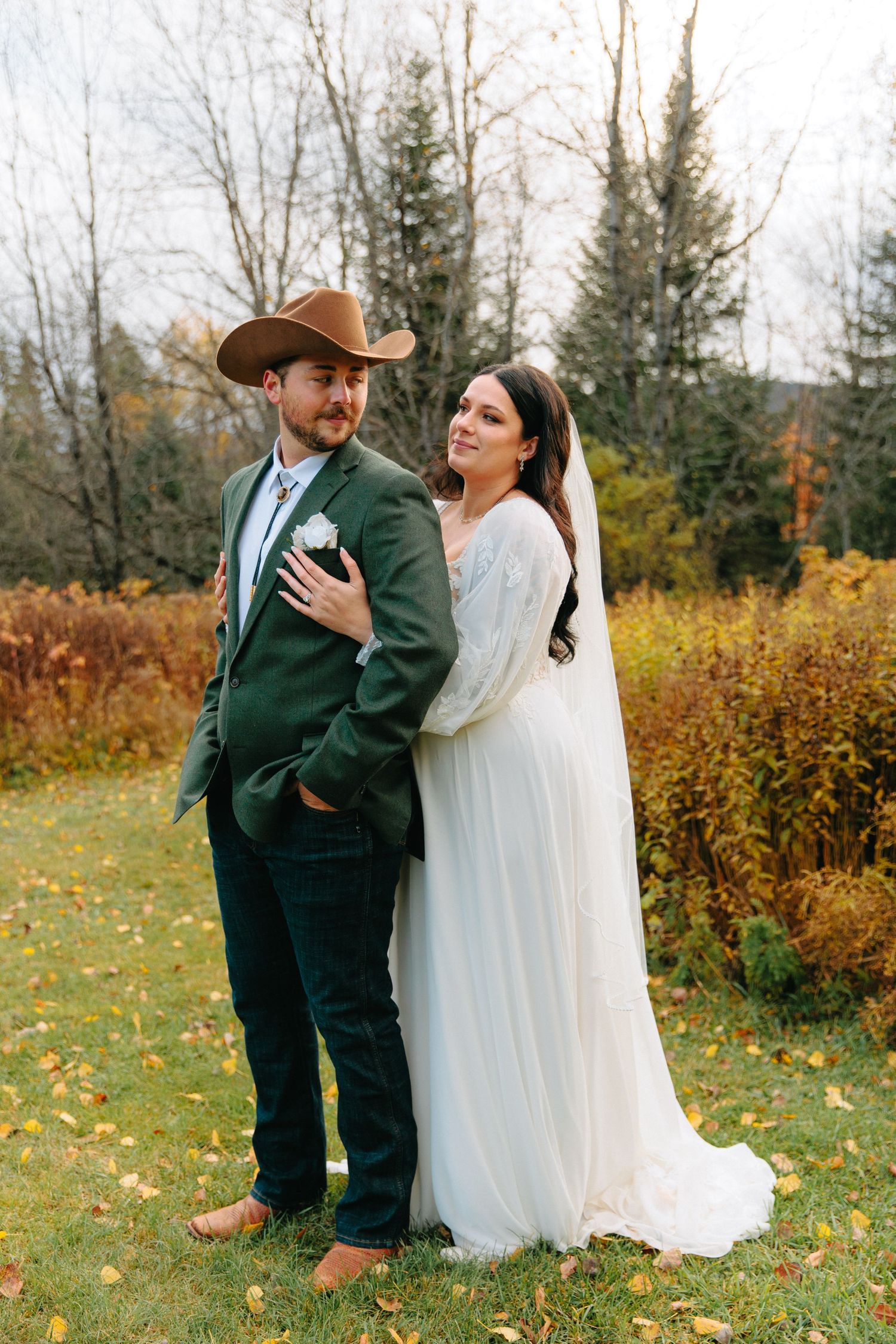 A couple shares a romantic moment in a fall meadow with evergreen trees and golden foliage in the background.