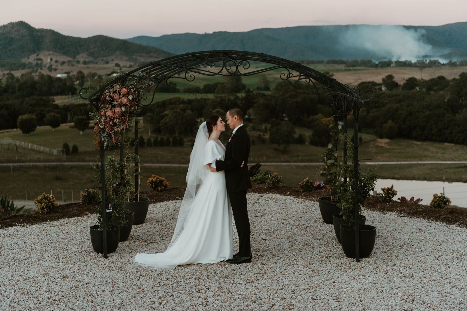 Bride and groom share a kiss under an iron arch with mountain views at a romantic outdoor wedding ceremony.