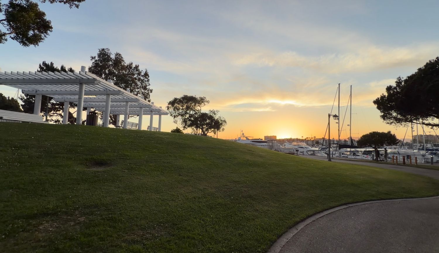 Sunset over a marina with sailboats, green lawn, and a white pergola structure at a waterfront park.