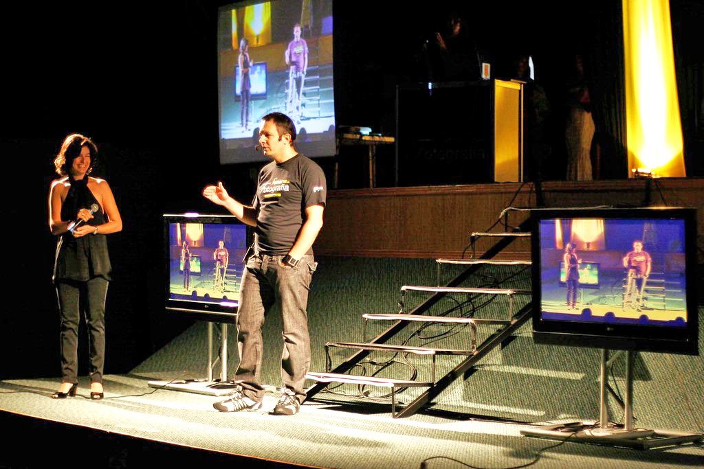 Two presenters on stage with monitors displaying fitness content during a live event presentation.