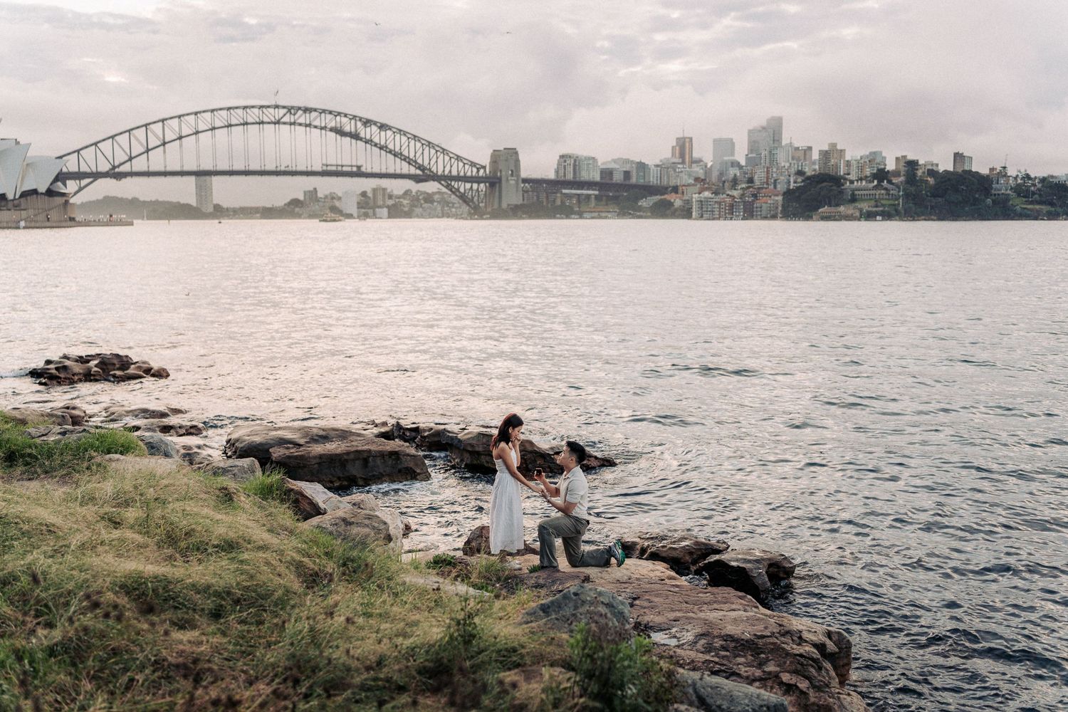 Couple sitting on rocky shoreline with Sydney Harbour Bridge and city skyline in the background.