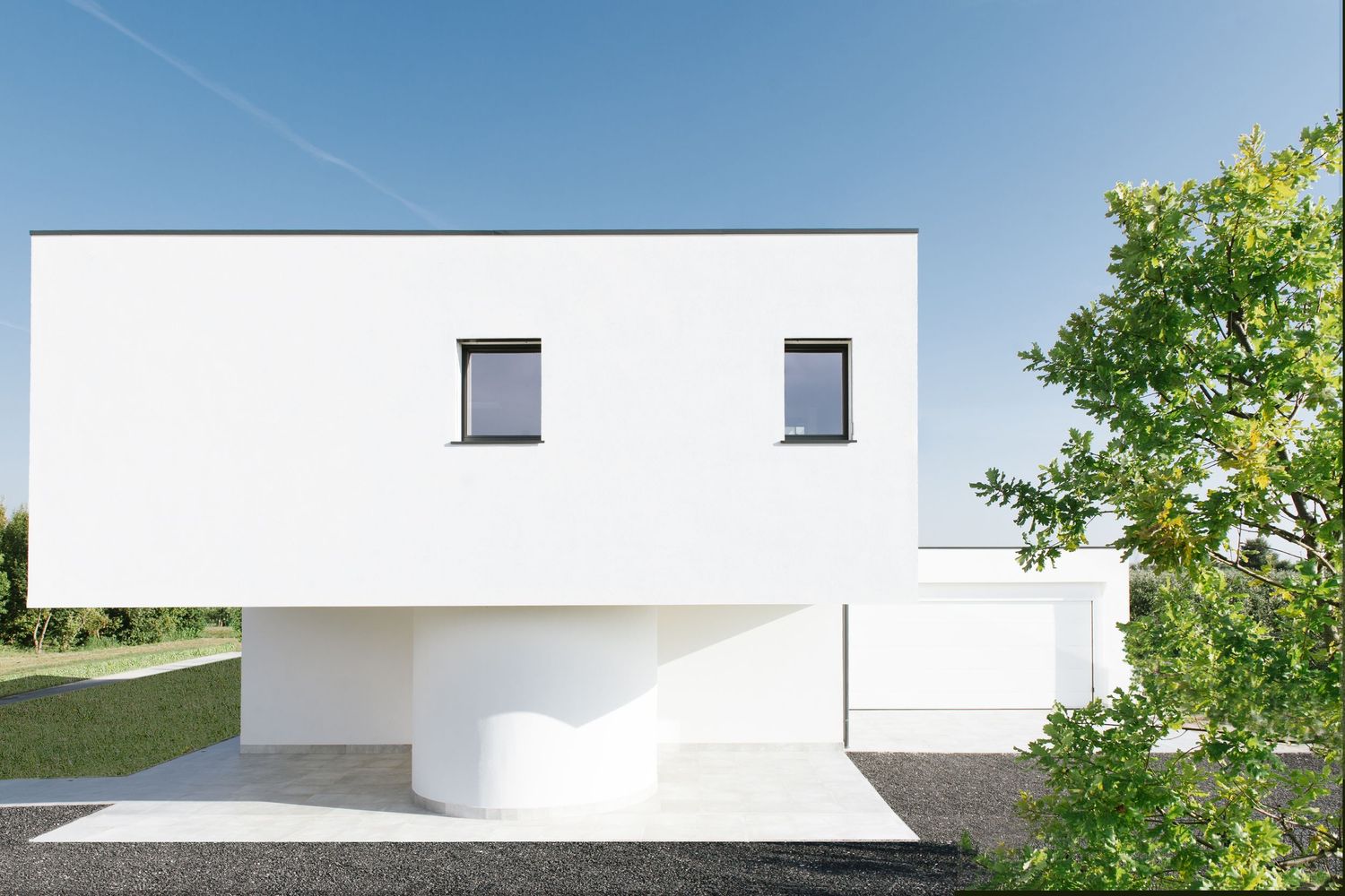 Modern minimalist white villa with large glass windows, circular accent, and lush green lawn under a clear blue sky.