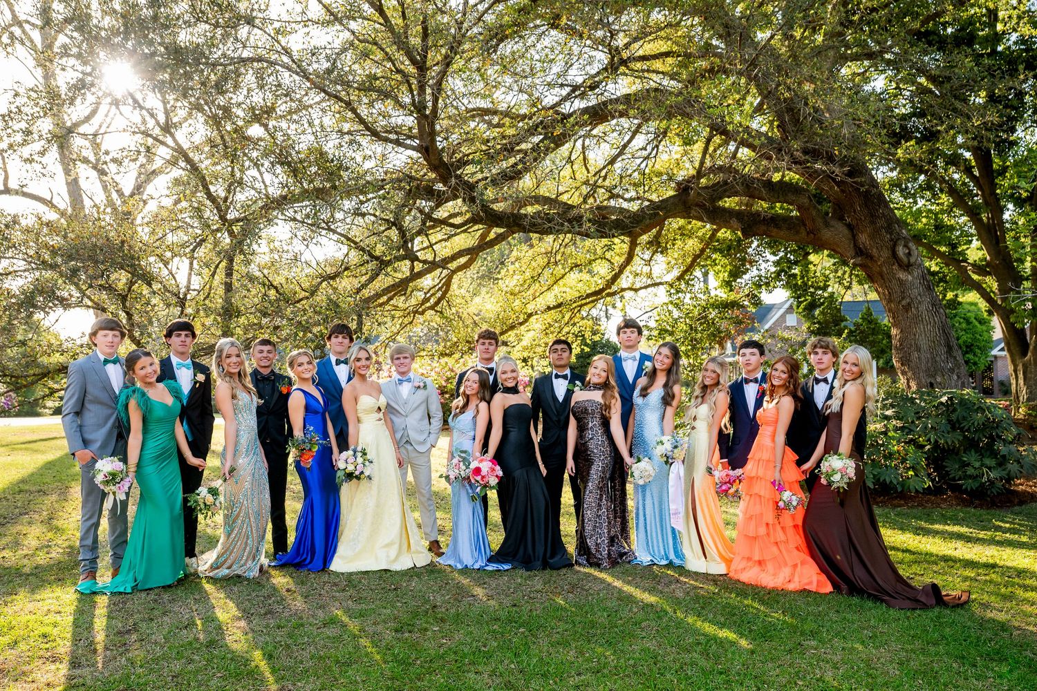 Large prom group in colorful formal gowns and suits posing outdoors under a sprawling oak tree on a sunny day.