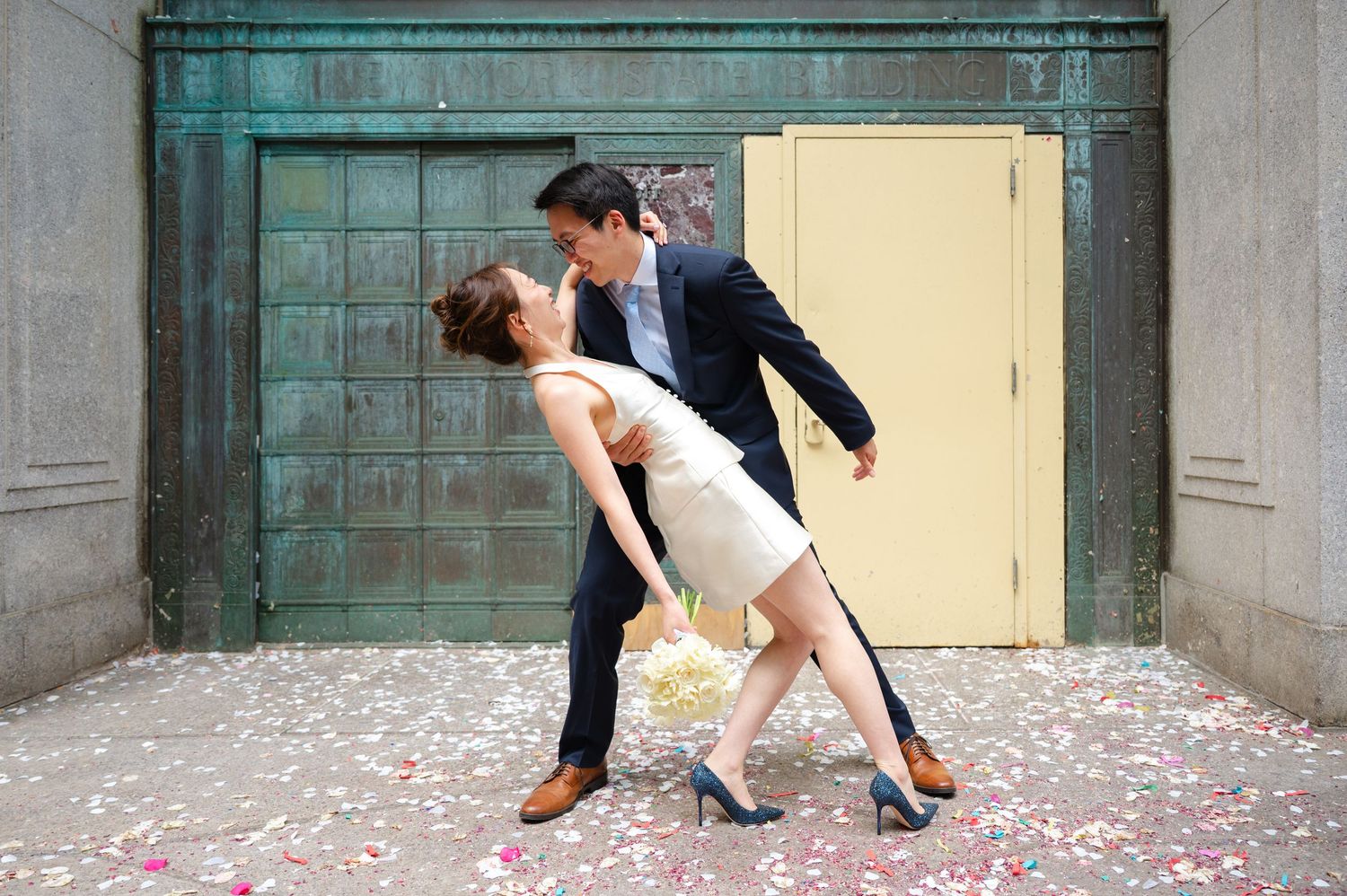Couple sharing a romantic dip kiss in white dress and suit, surrounded by colorful confetti petals on stone in front of NYC City Clerk.
