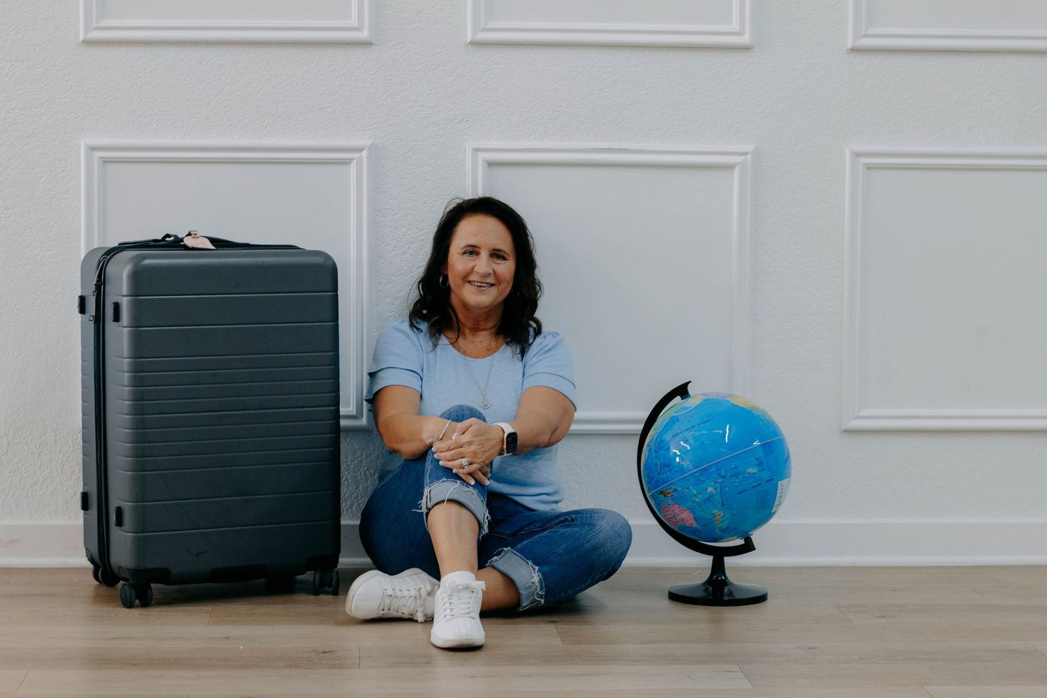 Woman sitting on floor between a suitcase and globe, smiling, wearing blue shirt and jeans, travel planning concept.