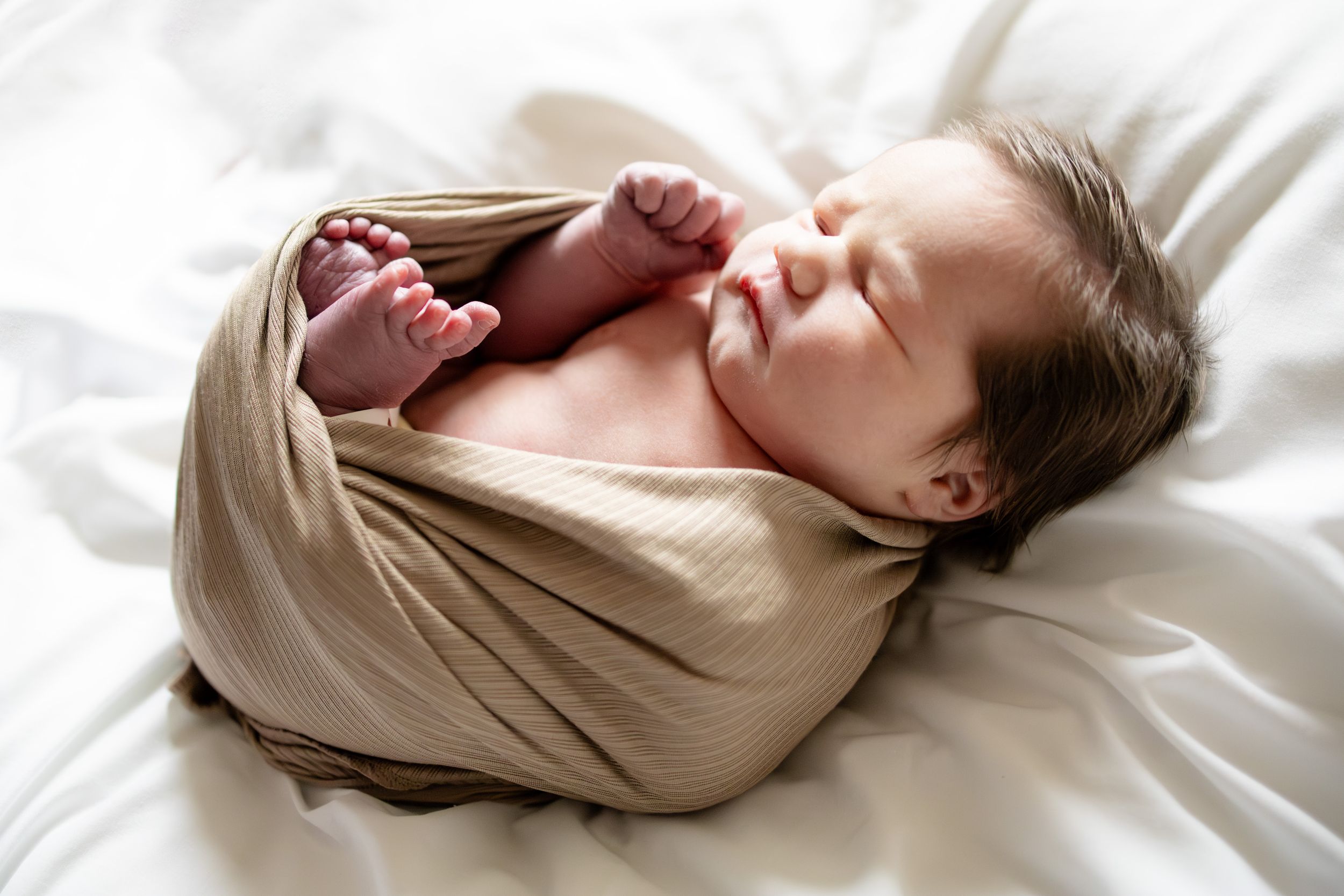 Peaceful newborn baby swaddled in a beige wrap, sleeping on white bedding with tiny feet peeking out.