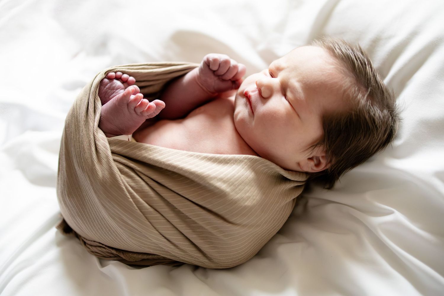 Peaceful newborn baby swaddled in a beige wrap, sleeping on white bedding with tiny feet peeking out.