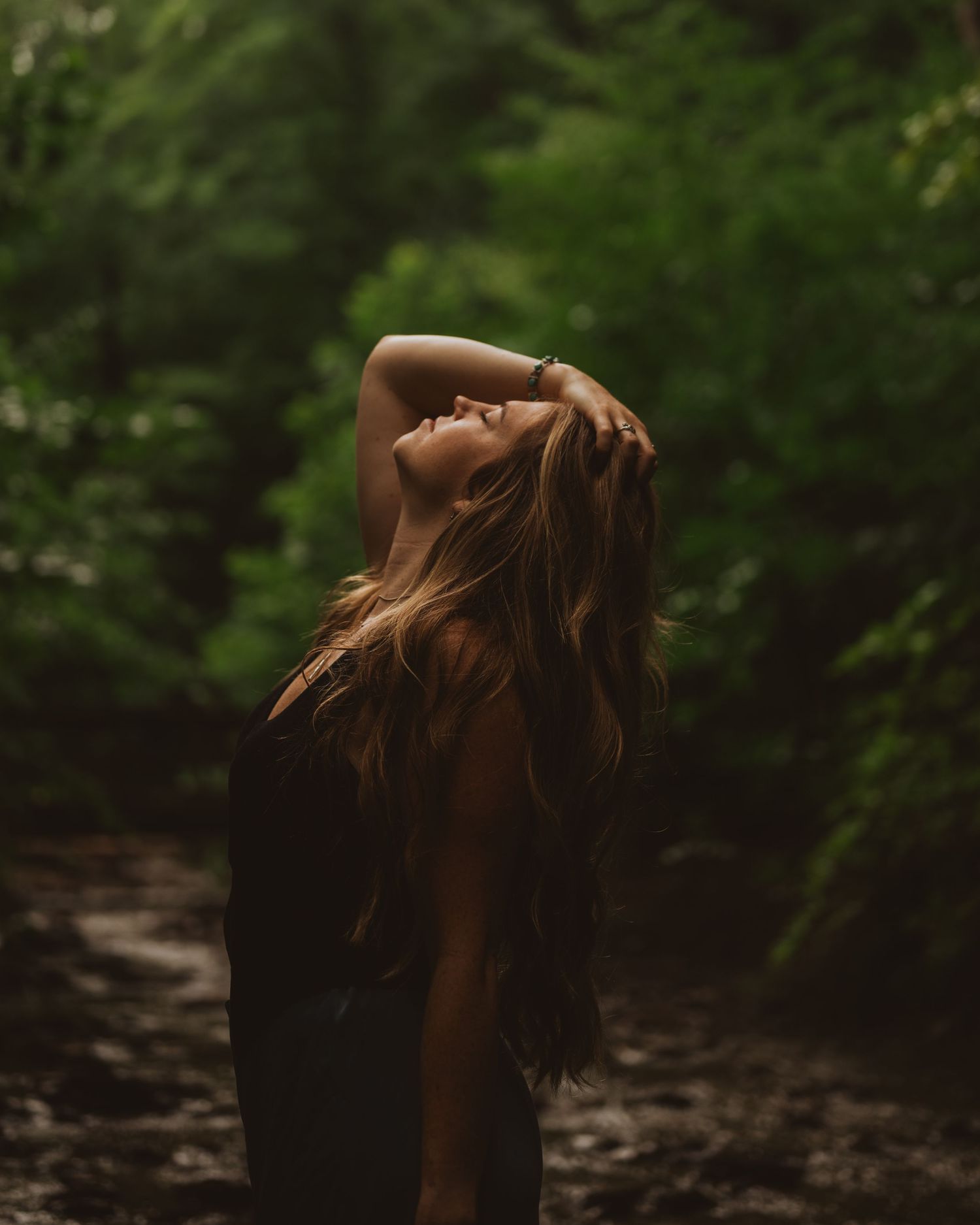 Woman with flowing brown hair throws her head back against a moody forest backdrop in dramatic silhouette.