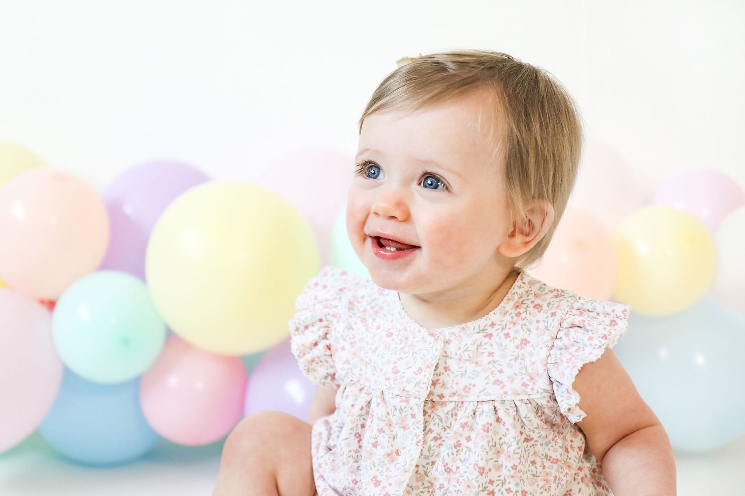 Baby in lace dress smiling with pastel balloons in background against white wall, during birthday shoot in Nottingham