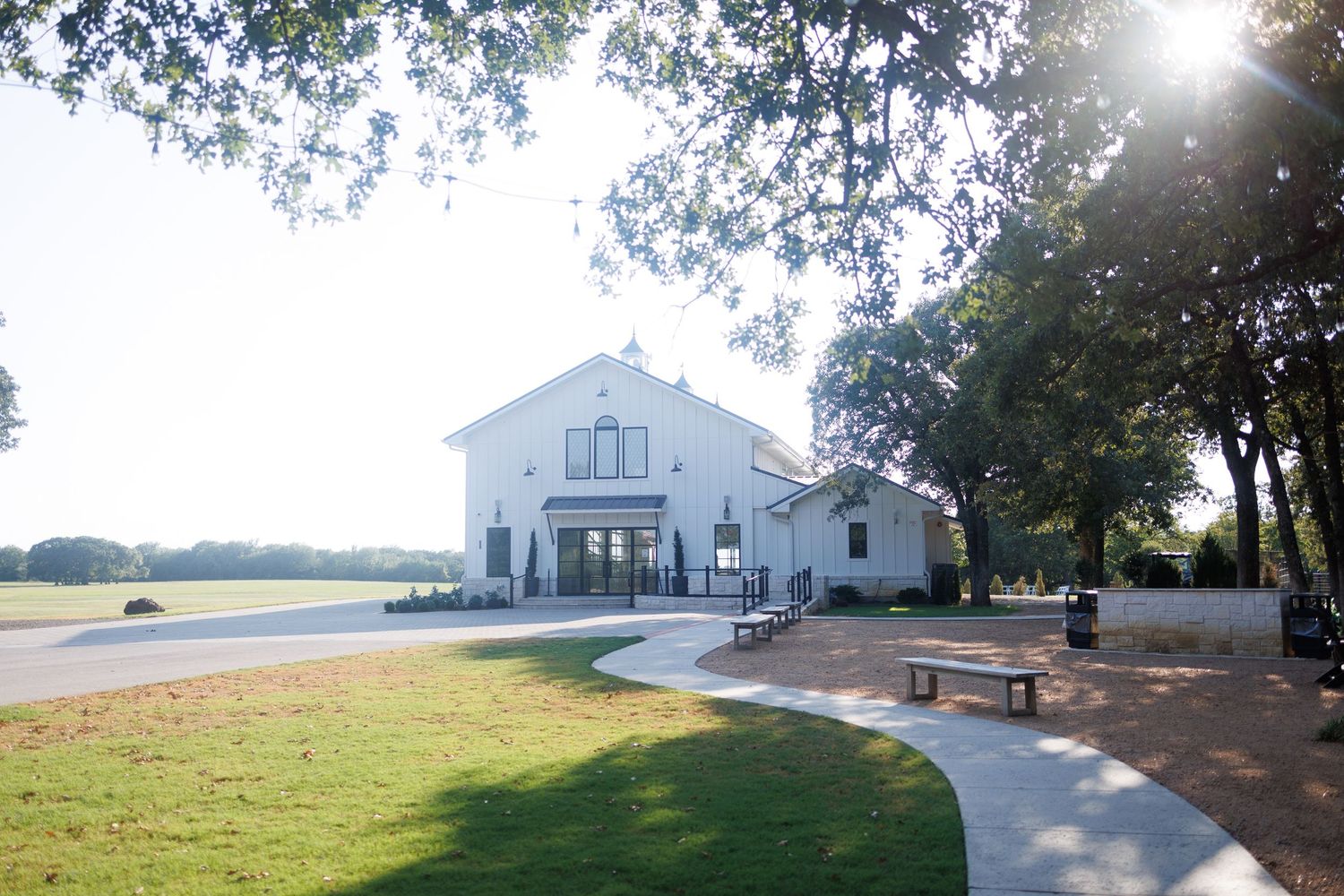 Bella Cavalli, a white rustic barn-style wedding venue near Dallas, with a curved walkway and picnic table under trees on a sunny day.