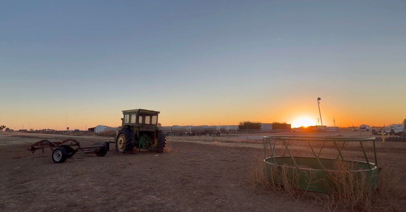 Farm tractor and hay bales silhouetted against a vibrant orange sunset over rural farmland.
