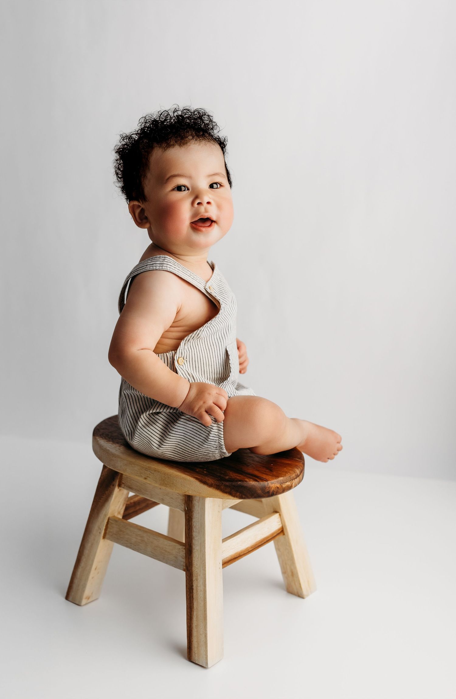 Baby sits on wooden stool wearing ribbed tank romper in minimalist white studio setting.