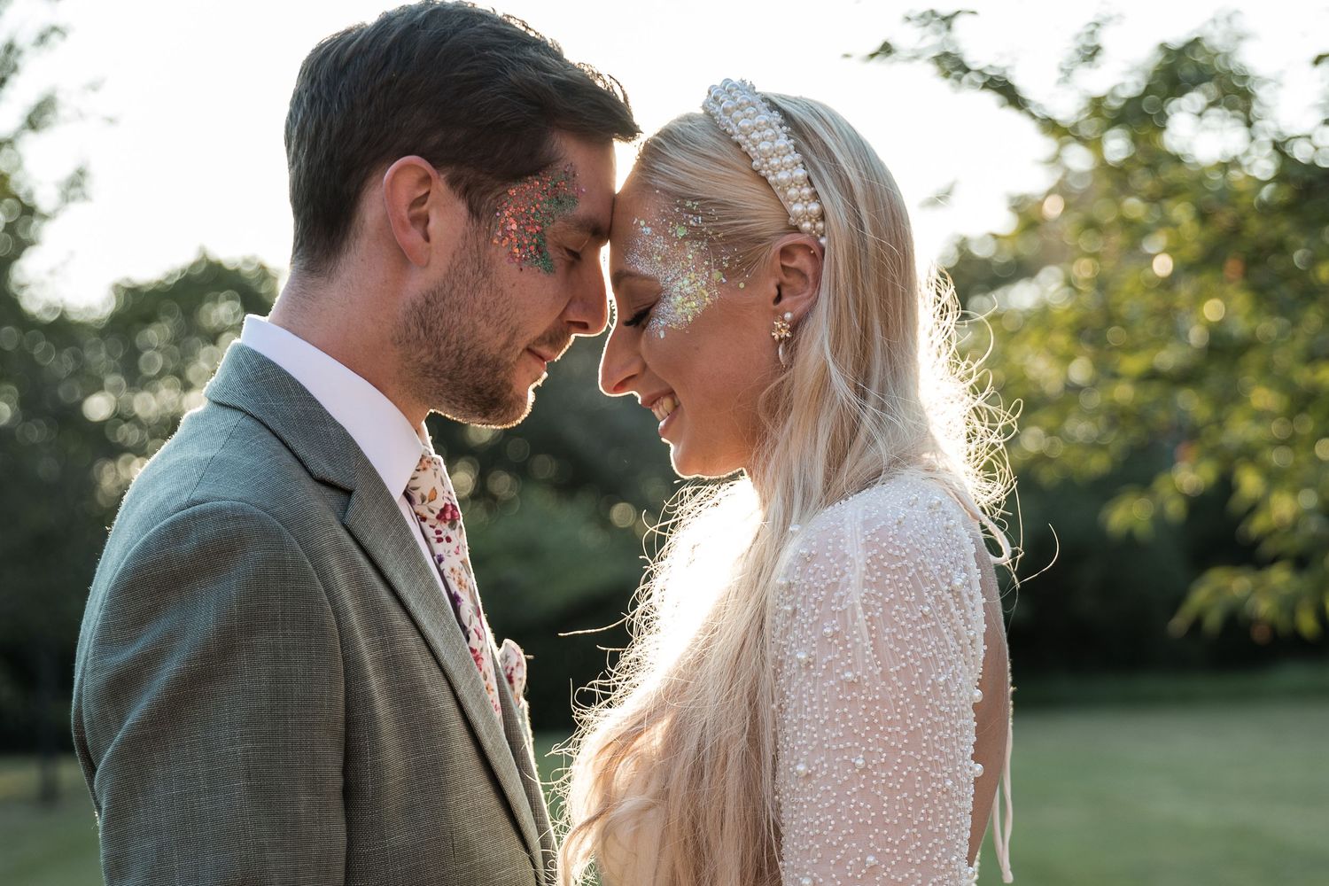 A couple in wedding attire share an intimate moment outdoors with sunlight filtering through trees in the background.