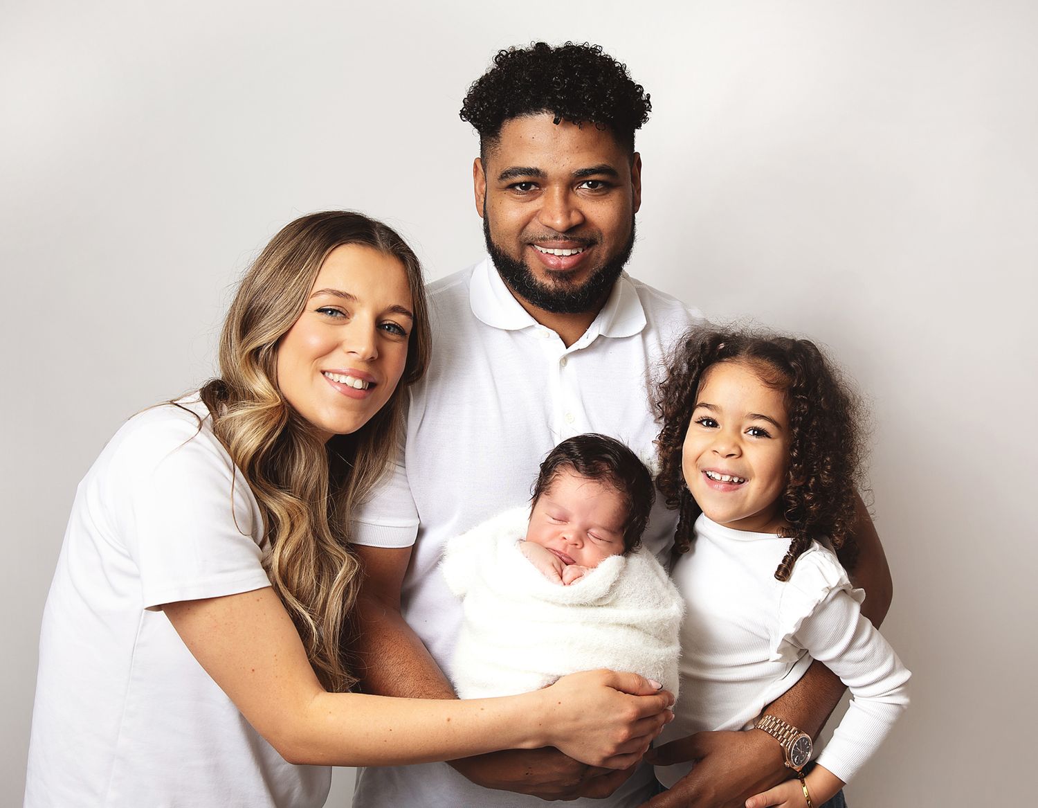 Family in white attire sitting together for portrait against light grey background. During a newborn family session in London