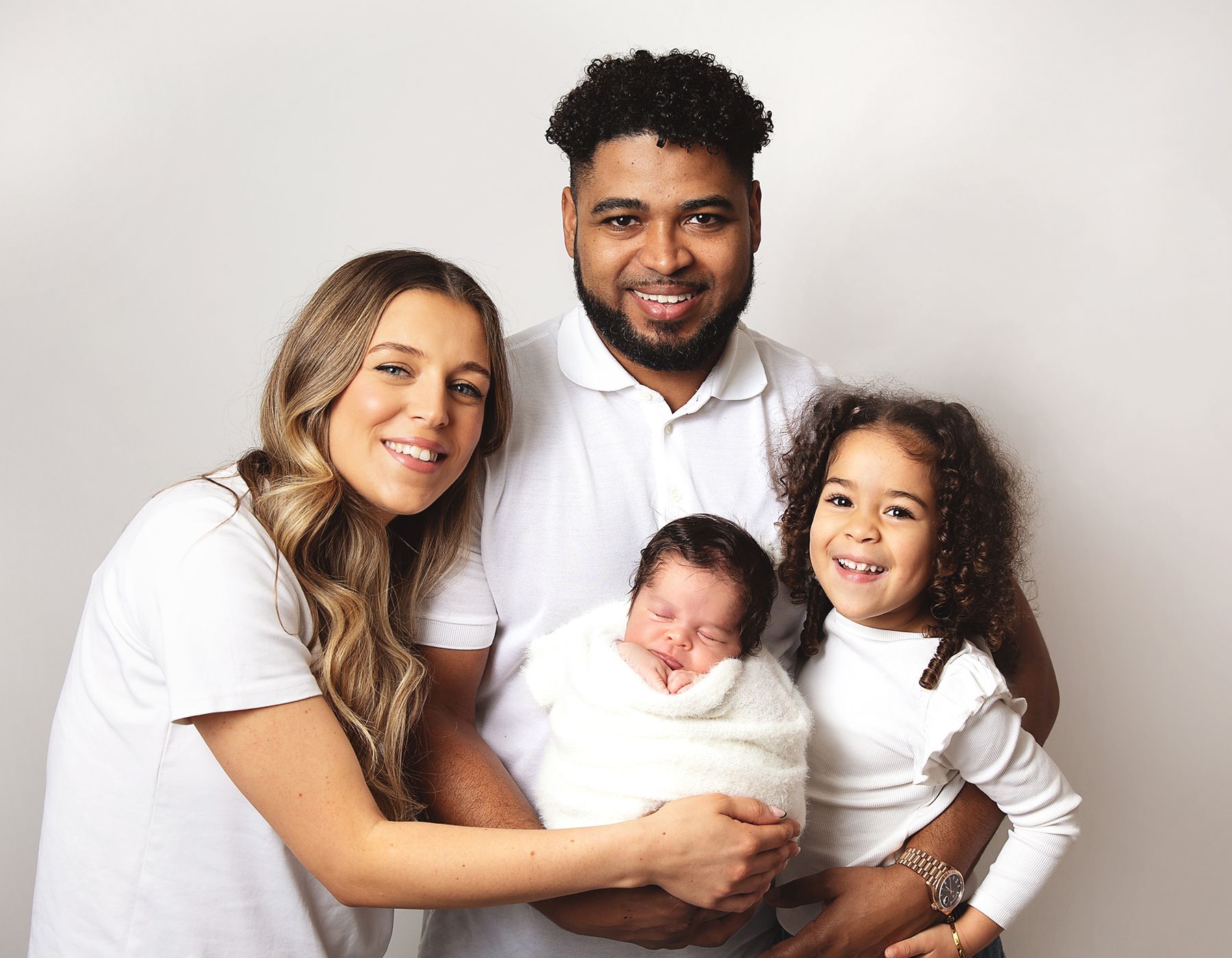 Family in white attire sitting together for portrait against light grey background. During a newborn family session in London
