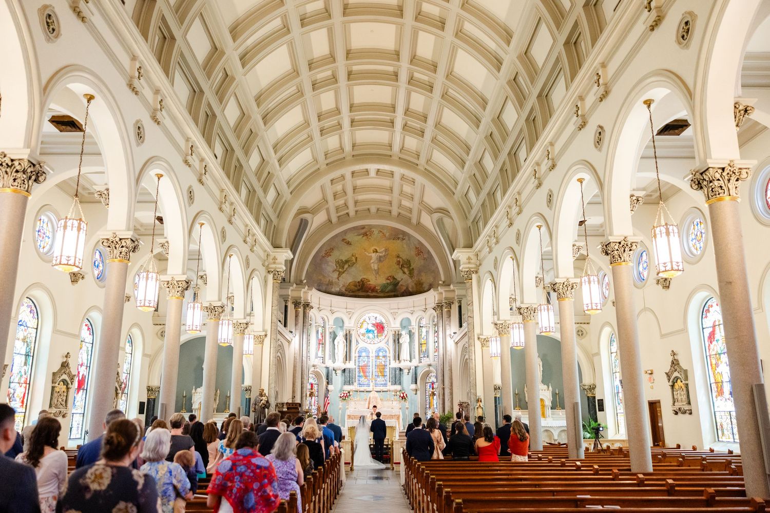 Wide angle view of ornate church interior with vaulted ceiling, gilded columns and crowded pews during ceremony.