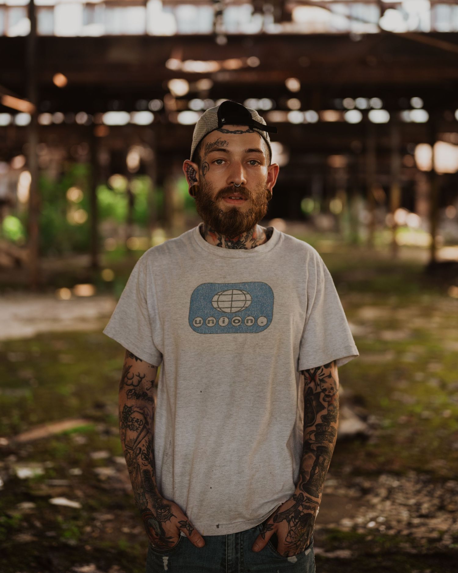 Heavily tattooed person wearing a backwards cap and gray t-shirt with a retro graphic logo stands outdoors.