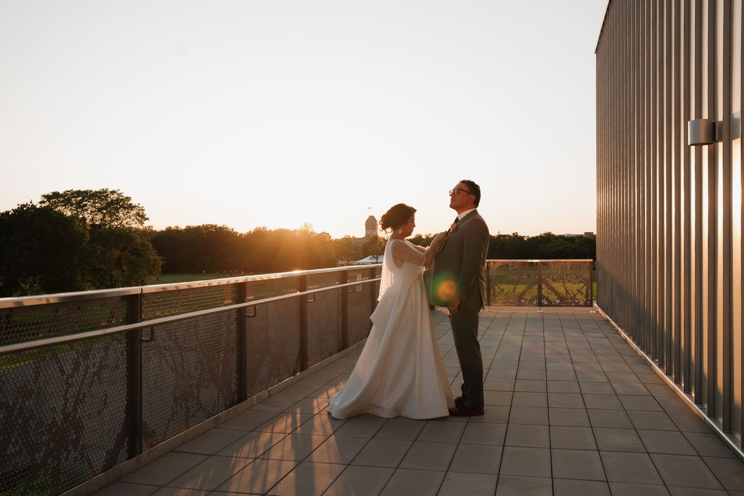 A couple stands together in a grassy field at sunset, the bride in a flowing white gown and groom in a gray suit.