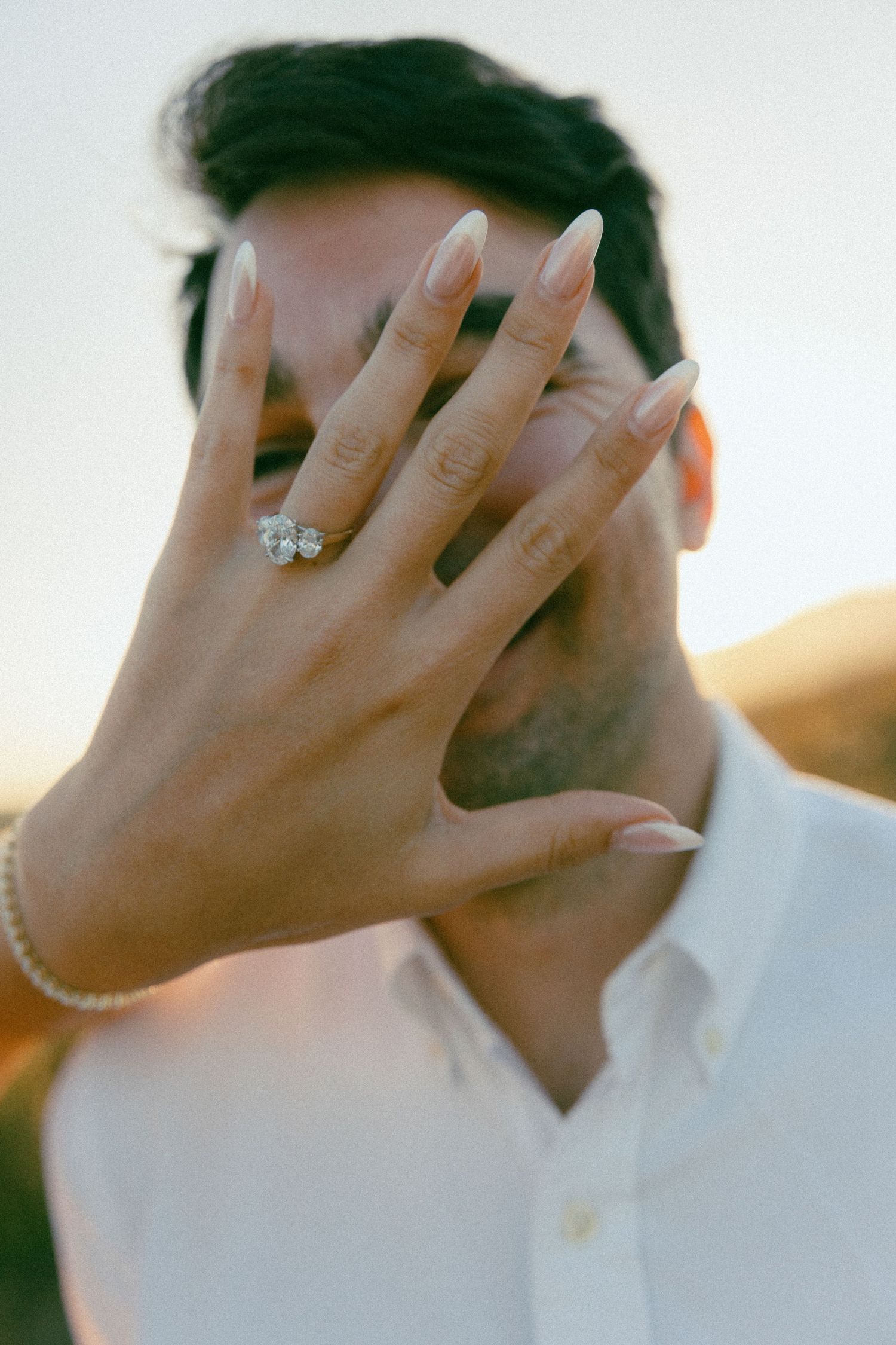 A hand wearing a sparkling engagement ring covers someone's face, with soft golden sunlight in the background.