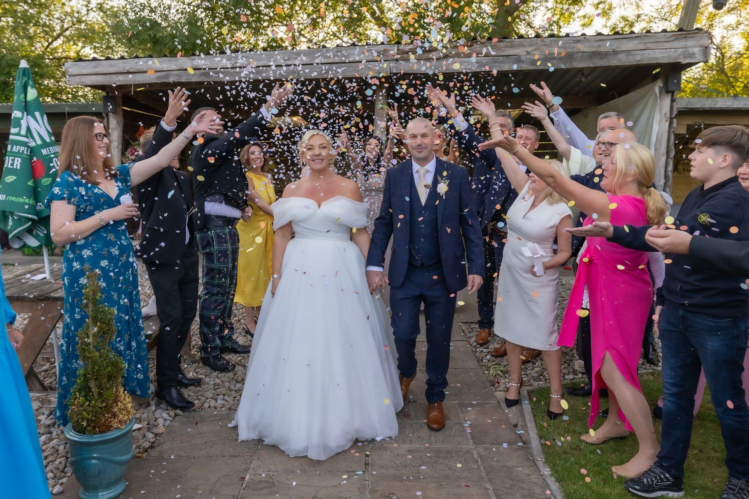 Wedding guests shower newlyweds with confetti outside rustic venue during daytime celebration.