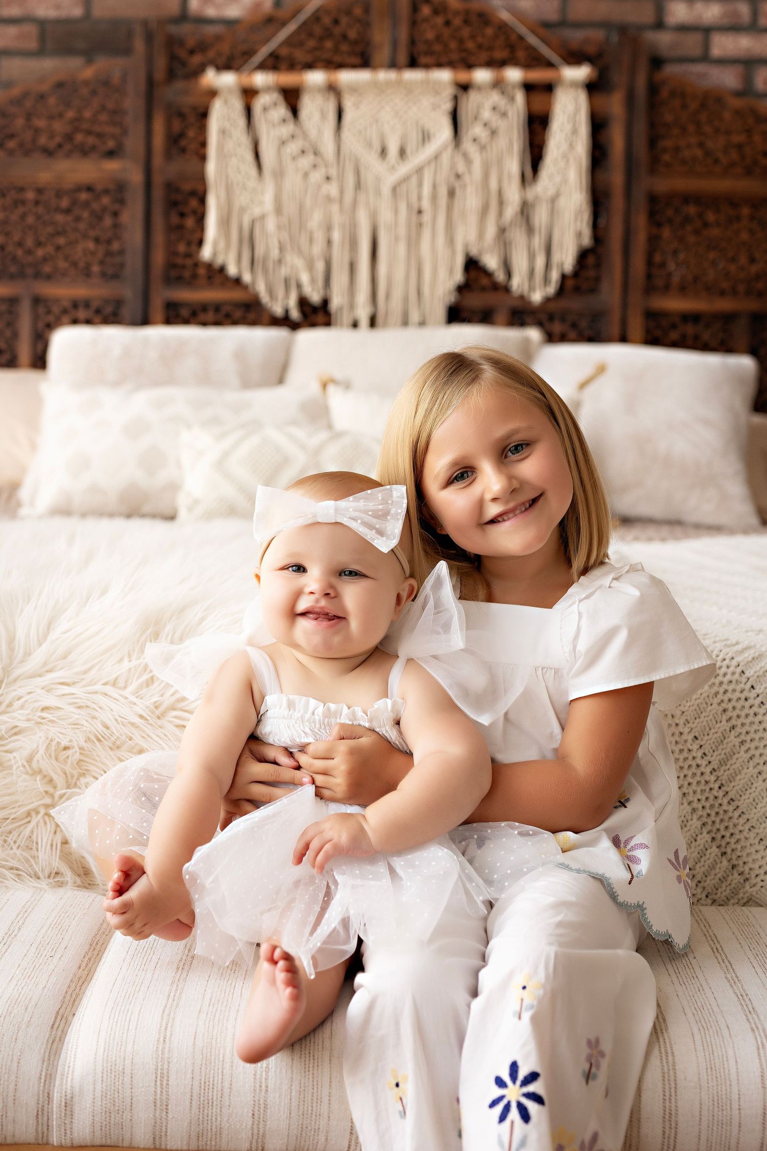 Two children in white outfits sitting together on a cozy bed with a macrame wall hanging above.