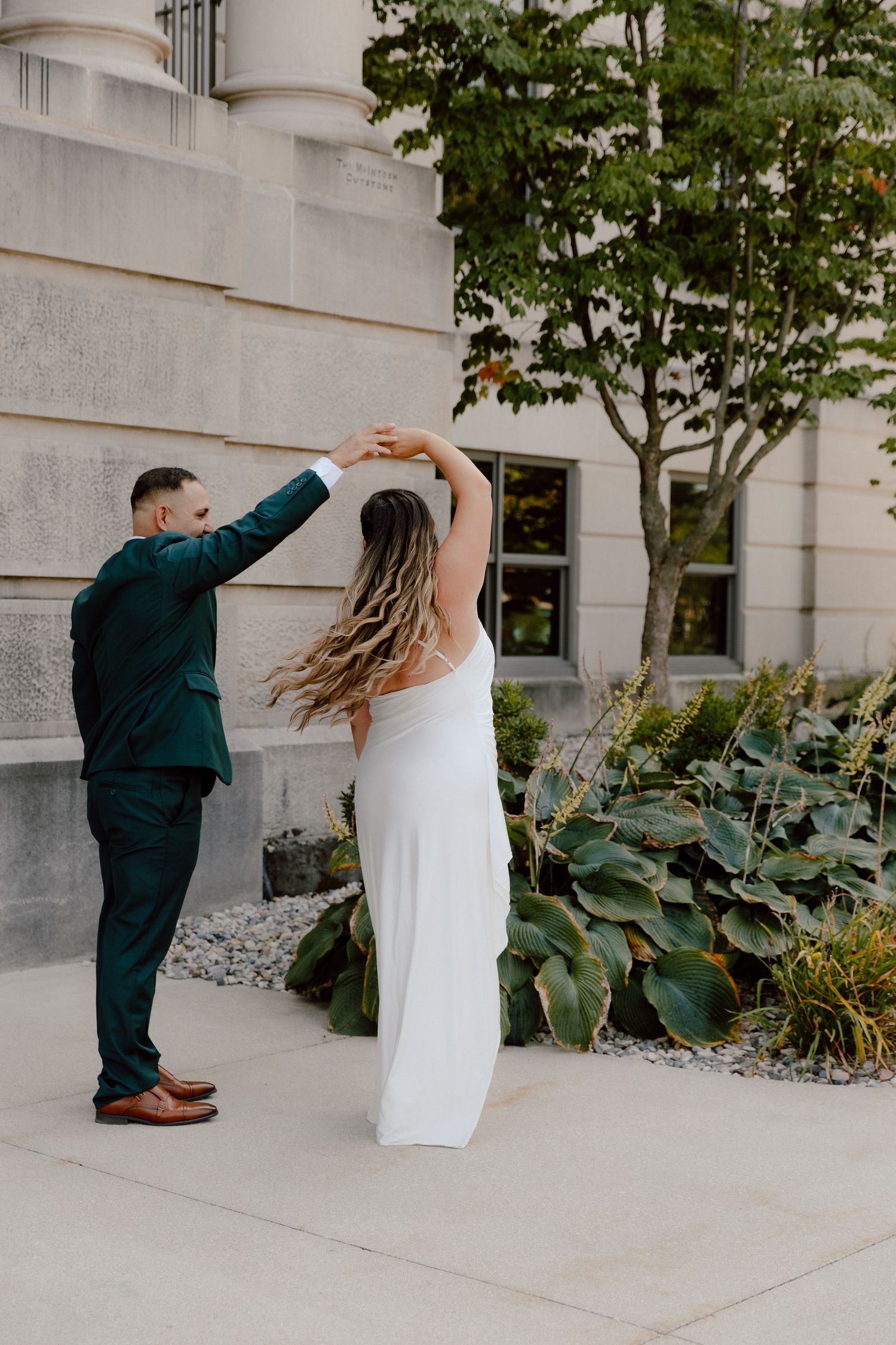 A couple dances together outside a building with elegant architecture and landscaping, the bride wearing a flowing white dress.