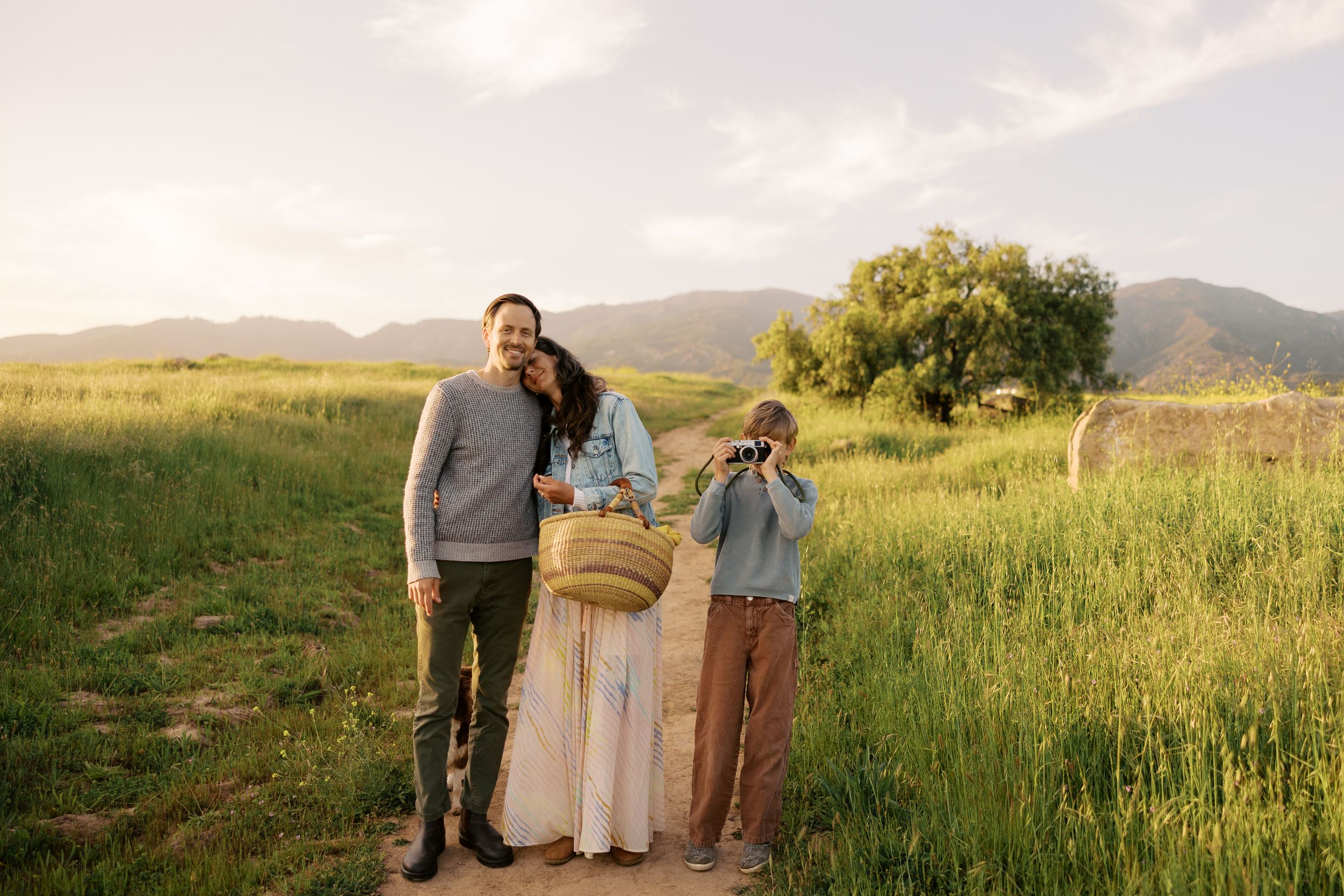 A family poses together in a scenic mountain meadow at golden hour with tall grass and trees in the background.