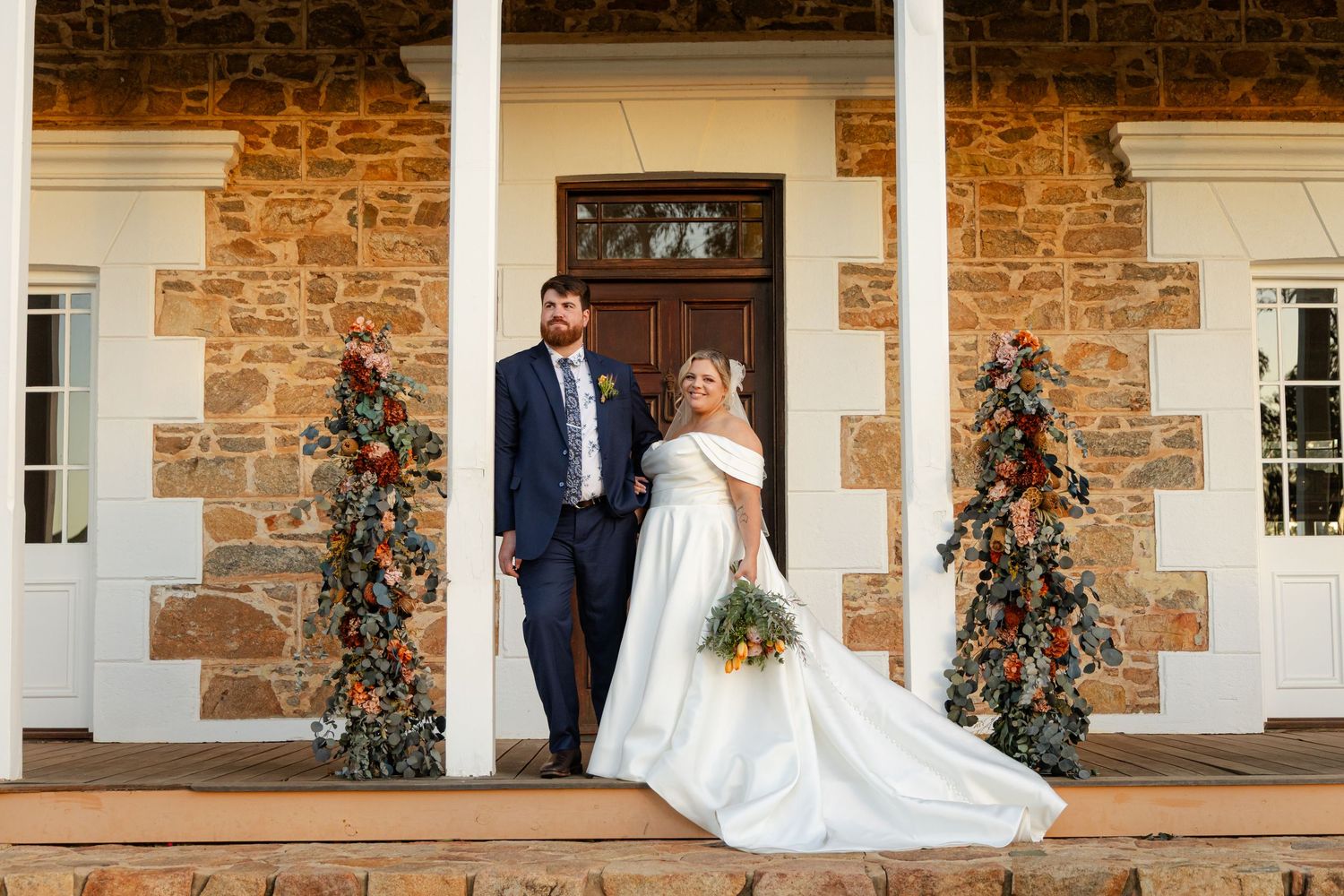 A couple stands on a rustic stone porch decorated with floral arrangements and greenery for their wedding day.