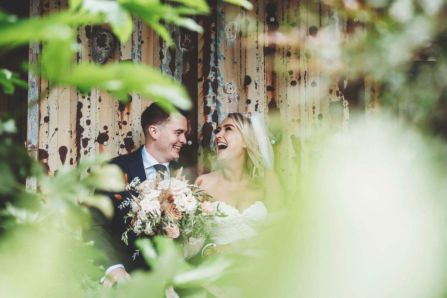 Newlywed couple in elegant wedding attire share a tender moment framed by lush green foliage in an outdoor setting.