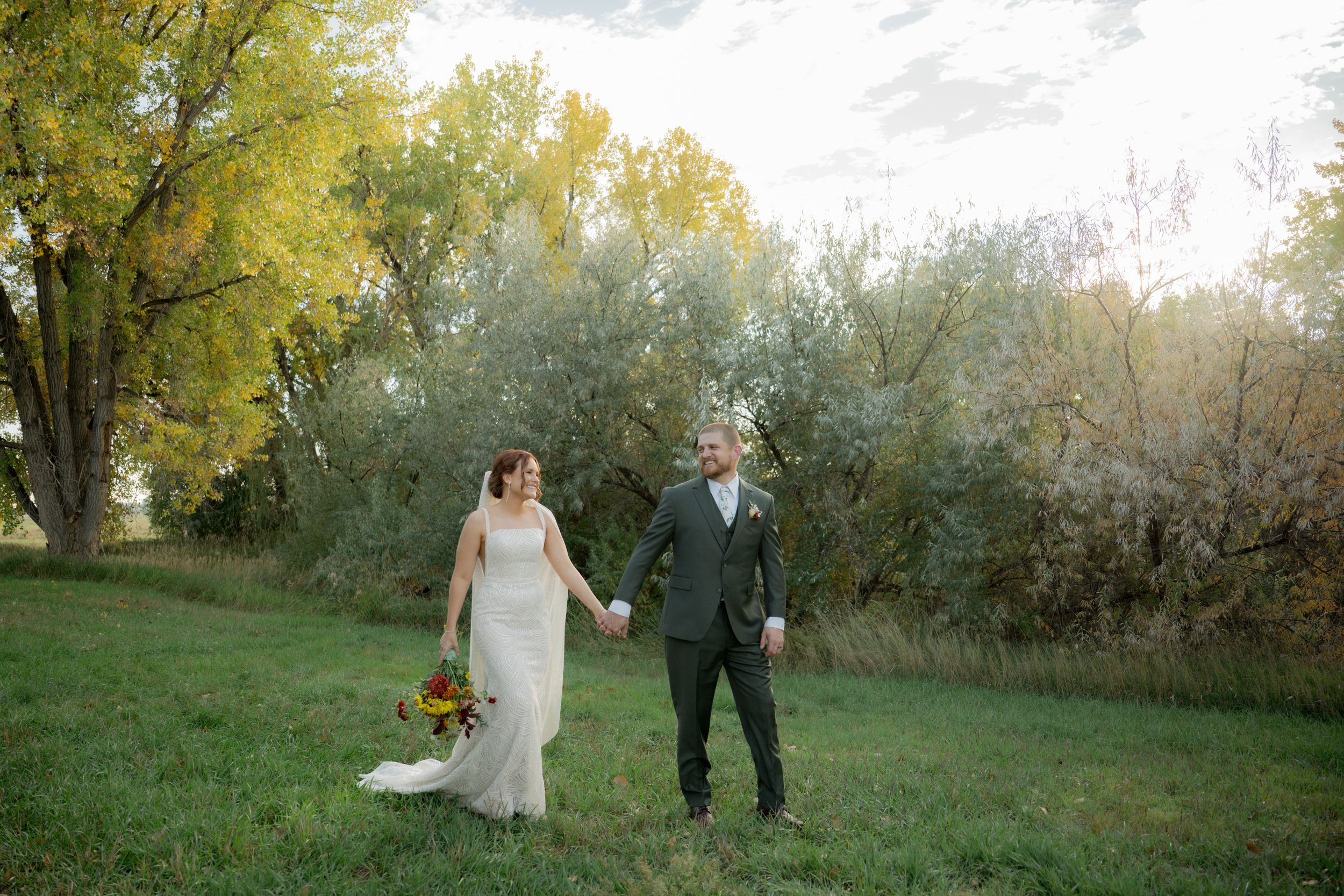 A couple walks hand in hand through a lush green field with autumn trees in the background on their wedding day.