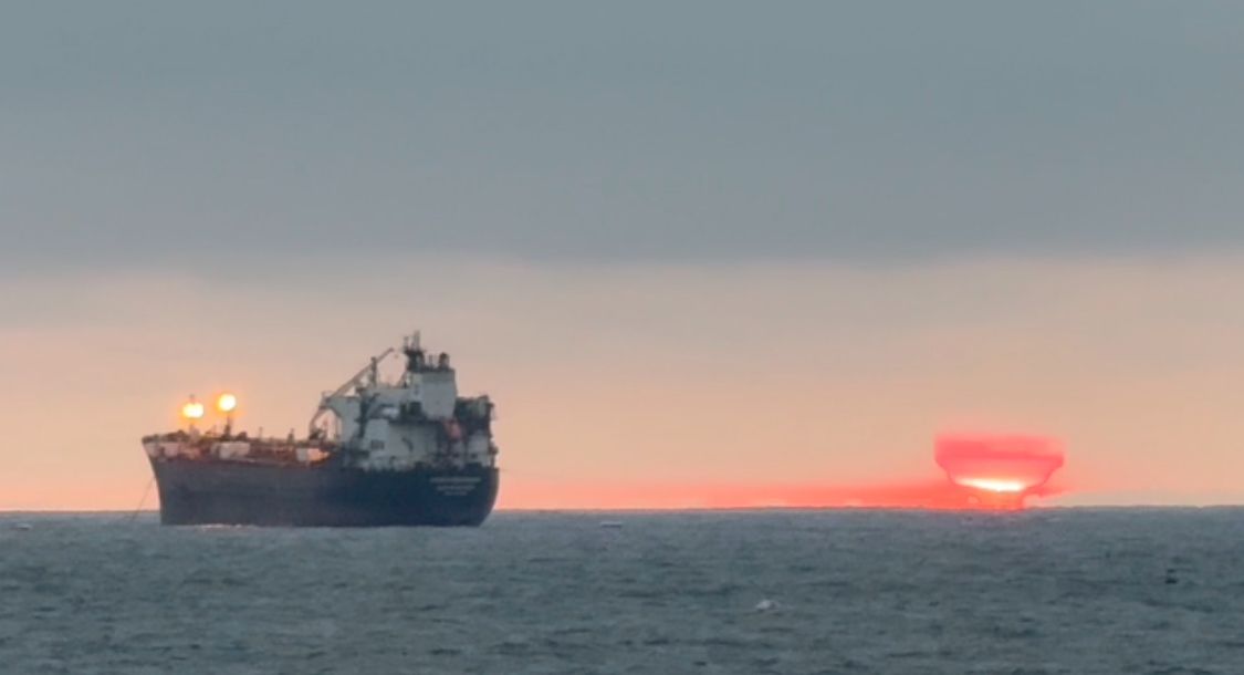 Silhouettes of cargo ships against a colorful orange and blue sunset horizon on the ocean.