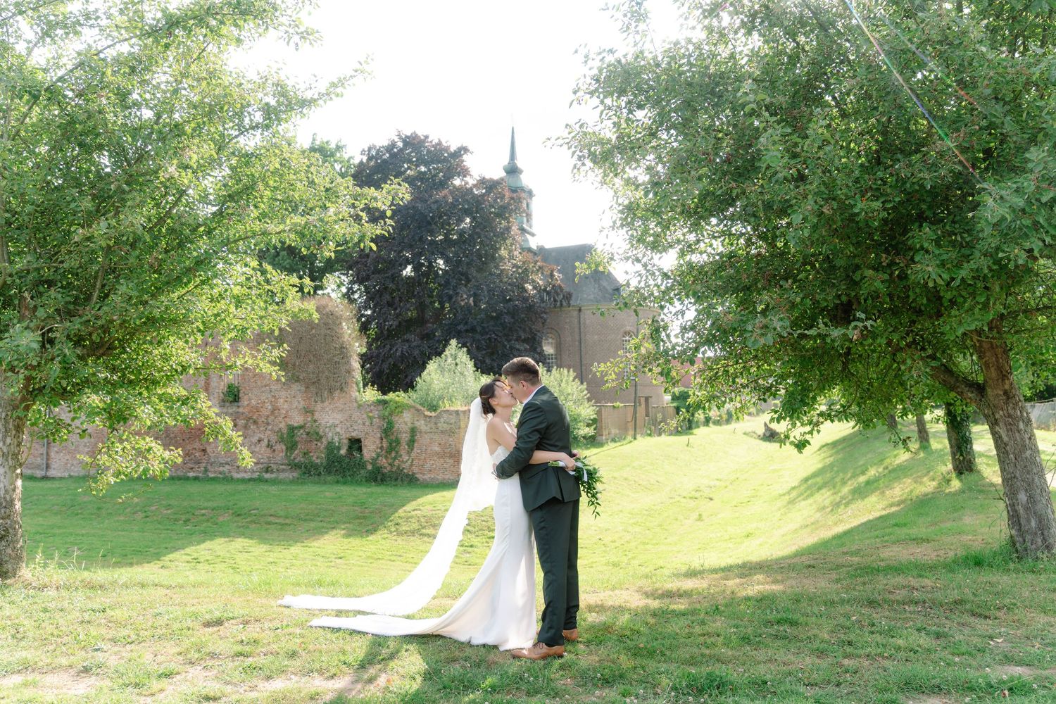 Wedding couple walks through lush green garden path with historic building in background.