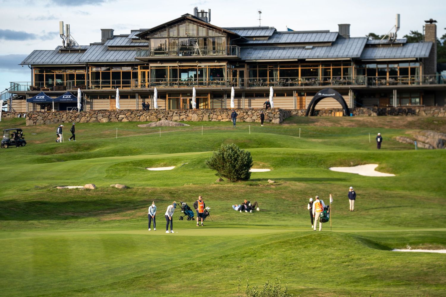 Panoramic view of a golf course with rustic clubhouse buildings and players scattered across the green fairways.