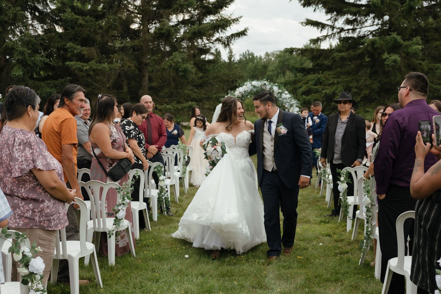 Panoramic view of an outdoor wedding ceremony with white chairs arranged on a grassy lawn surrounded by evergreen trees.