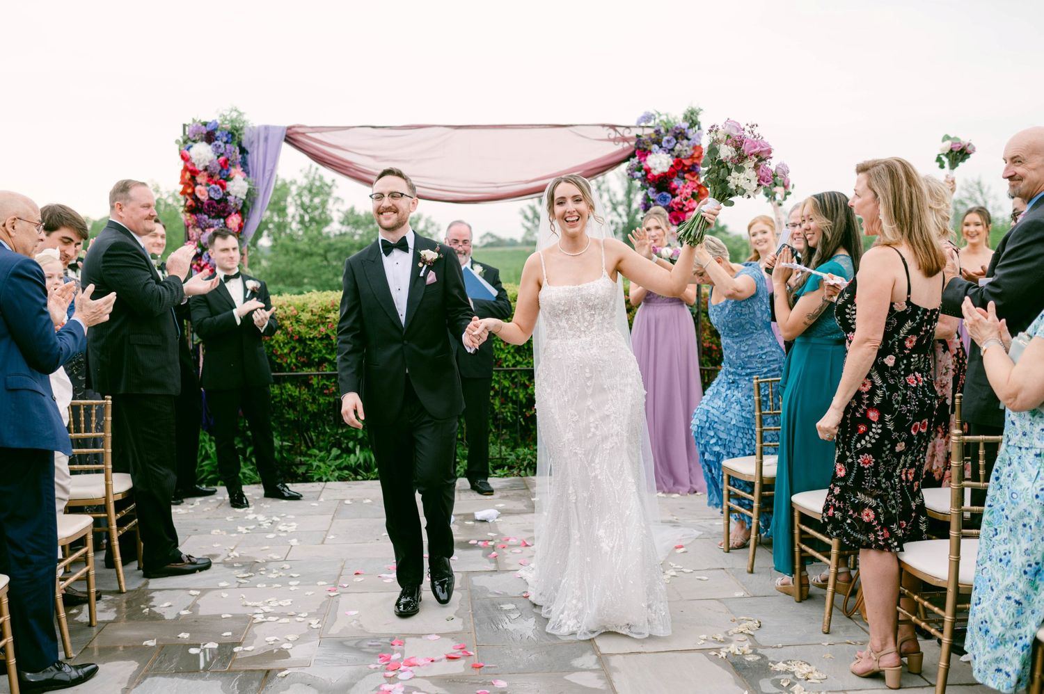 A couple celebrates at an outdoor wedding reception surrounded by cheering guests and wooden chairs against a scenic backdrop.