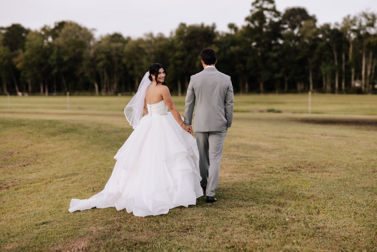 A couple in wedding attire walking through a grassy field together at sunset.