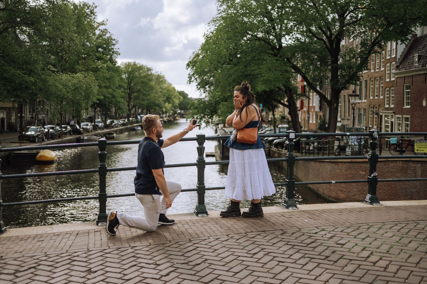Man kneeling and proposing on a quiet bridge over Amsterdam canal, emotional reaction from his partner.