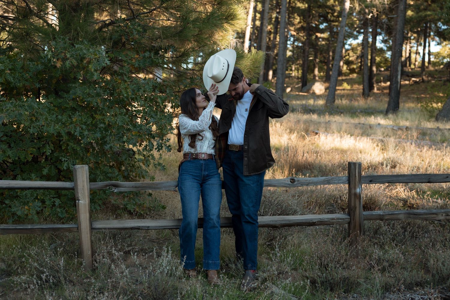 A couple in casual attire stands together on a wooden boardwalk surrounded by tall pine trees at sunset.