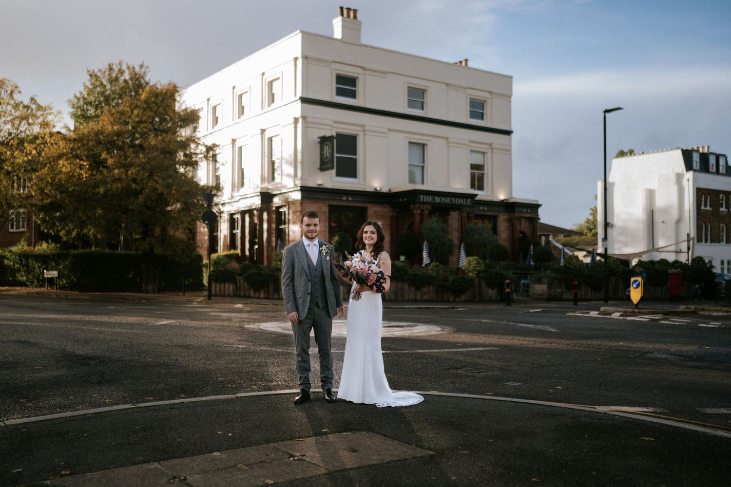 Couple in wedding attire poses on quiet urban street corner with historic white buildings in background.