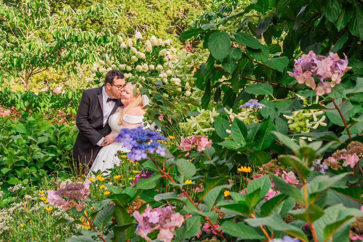 A romantic wedding kiss between a couple surrounded by guests in a lush garden setting during an outdoor ceremony.